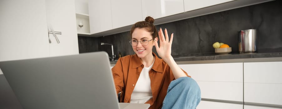 Cheerful young woman in glasses, saying hello, waving hand at laptop, talking to friend online, video chatting, sitting in kitchen with computer.