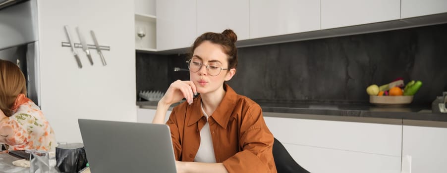 Portrait of young woman, student studying remote, connects to online lesson on laptop, sitting at home and freelancing, working from her kitchen.