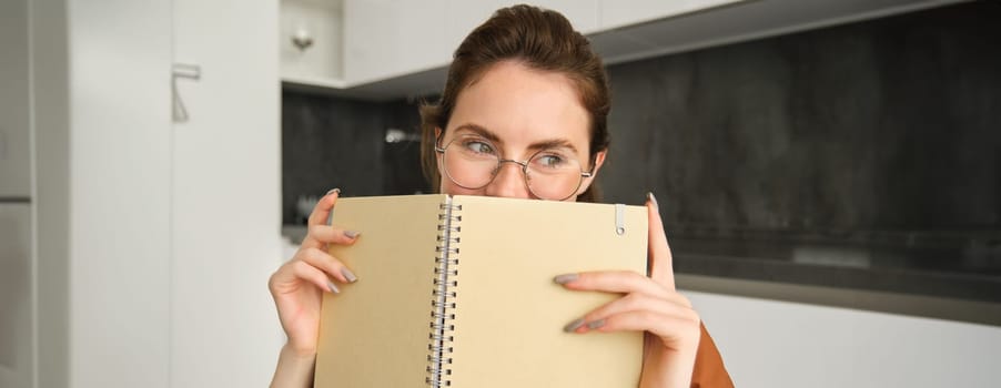 Close up portrait of cute smiling woman in glasses, sitting in kitchen at home, hiding face behind notebook and looking aside.