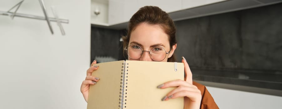 Close up portrait of cute brunette woman in glasses, sitting at home, smiling, hiding face behind yellow notebook.