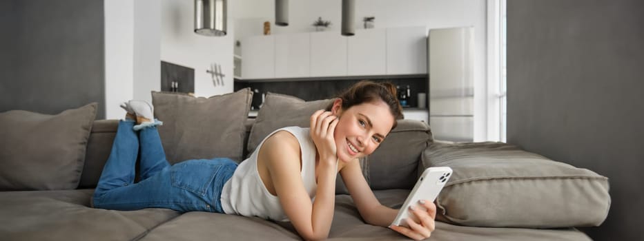 Portrait of young, beautiful young woman at home, resting on couch, holding smartphone, using mobile phone, looking at camera with happy smile.