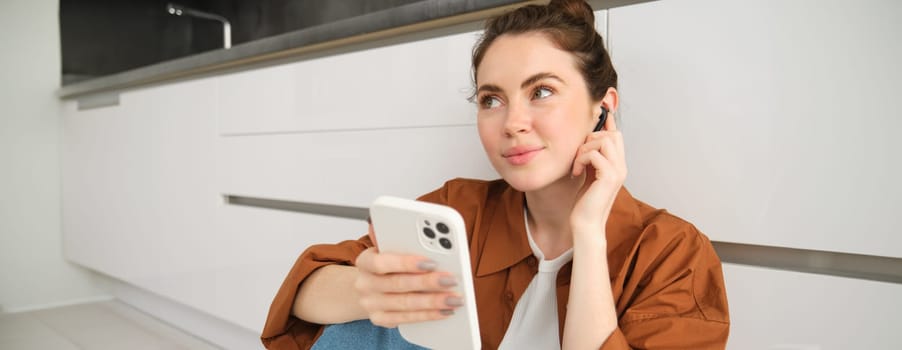 Portrait of young modern woman in wireless headphones, sitting on kitchen floor, using smartphone, making playlist with favourite songs, listens to music.