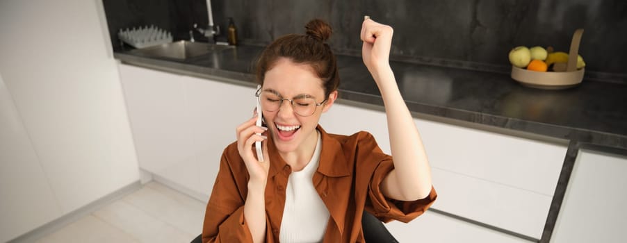 Portrait of woman receiving great news over the phone. Girl talking on mobile telephone and celebrating, laughing and making fist pump, dancing on her chair in kitchen.
