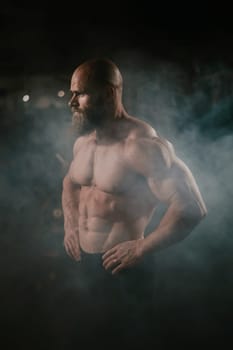 A muscular bald man poses shirtless in the dark amid smoke. A bodybuilder shows off his form in the gym