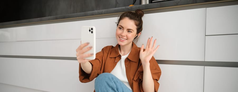 Portrait of happy smiling woman video chats, sits on kitchen floor at home, talking to friend on social media app using smartphone.