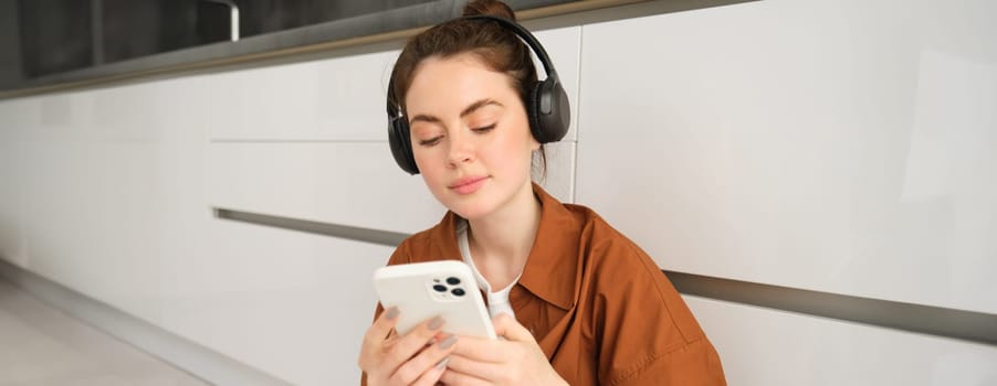 Young modern woman sending a message on smartphone, sits at home in wireless headphones, listens music, watches video on mobile app.