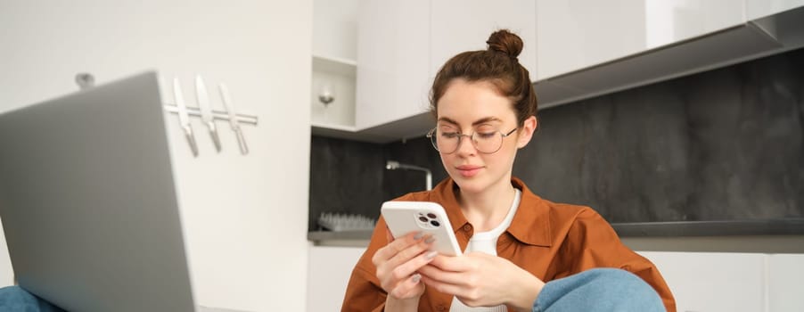 Portrait of young woman in glasses, checking her phone, working on remote from home, looking at smartphone, sitting in kitchen with laptop.
