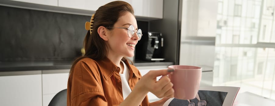 Close up portrait of young beautiful woman in glasses, spending time at home, sitting with cup of coffee, warming up with fresh mug of tea, resting in kitchen.