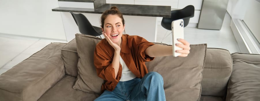 Photo of smiling young woman sitting on couch, posing on sofa in living room, taking selfies for social media.