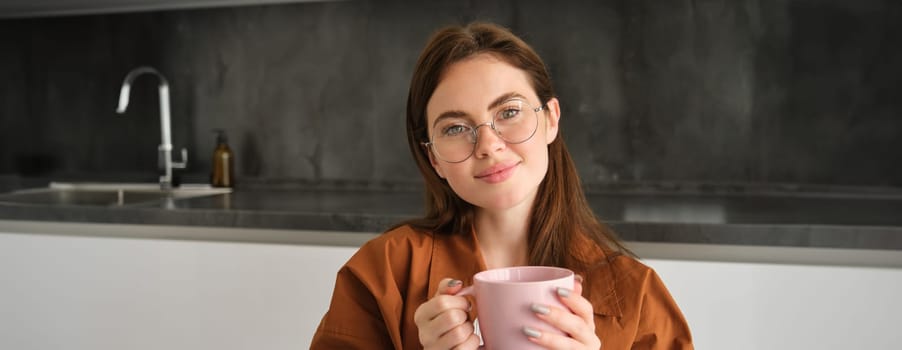 Portrait of lovely, tender young woman in glasses, sits at home with cup of tea. Girl in kitchen, enjoys drinking aromatic coffee.
