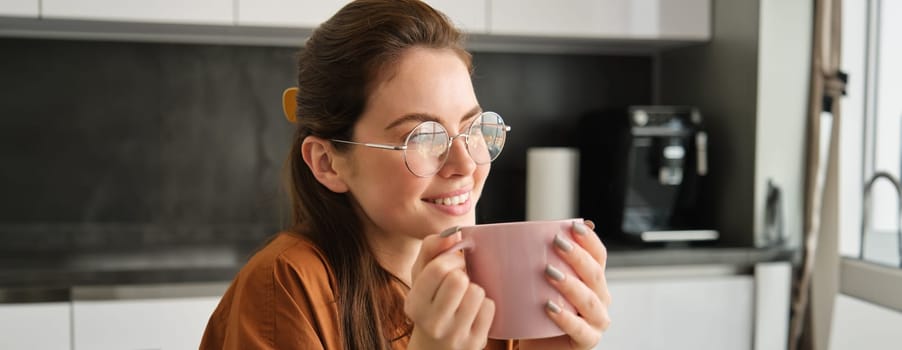 Portrait of happy woman, relaxing with cup of coffee, looking away, sitting in kitchen with pink mug, drinking herbal tea.