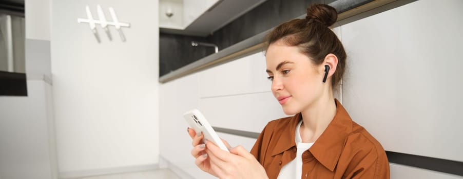 Close up portrait of young modern woman sits on kitchen floor, looking at her smartphone, watching video or tv series on streaming service app, listening music in wireless headphones.
