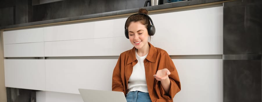 Portrait of young smiling woman explains something over the video call, sits in headphones with laptop, works from home.