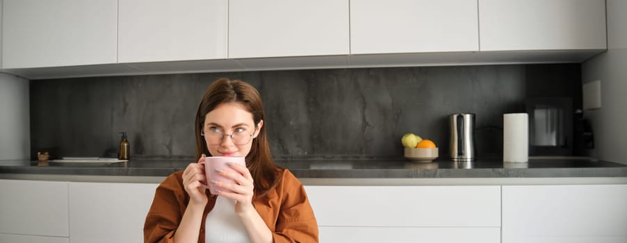 Portrait of brunette woman in kitchen, holding cup of tea, drinking aromatic coffee at home and enjoying calm, cosy day off.