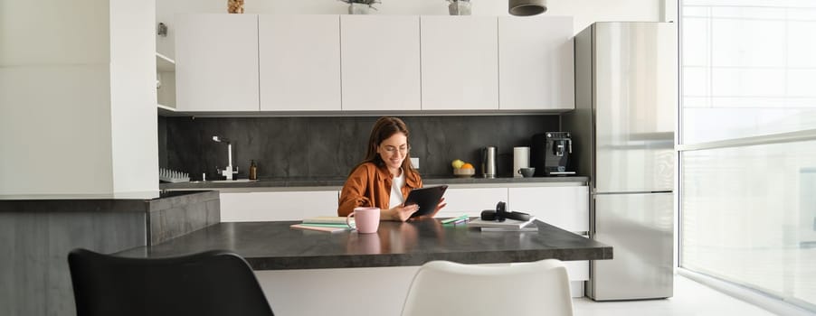 Portrait of young woman, student sitting in kitchen with tablet and notebook, reading news online, studying remotely, e-learning at home.