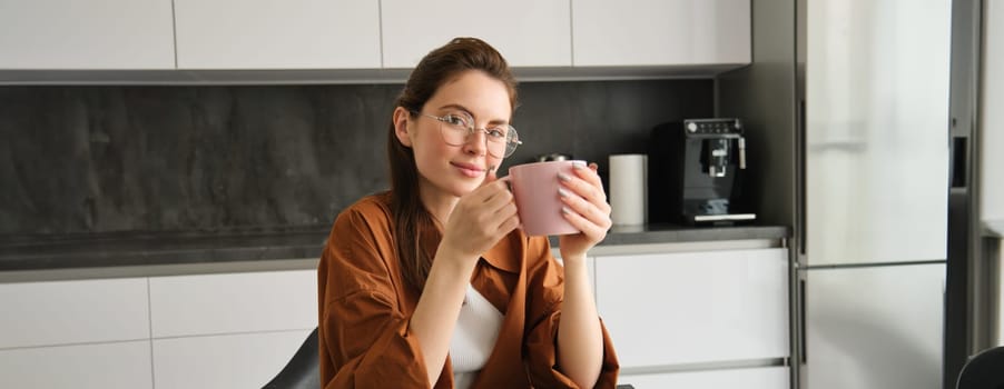 Portrait of beautiful brunette woman, sitting in kitchen, taking a break for cup of coffee. Girl drinks tea at home.