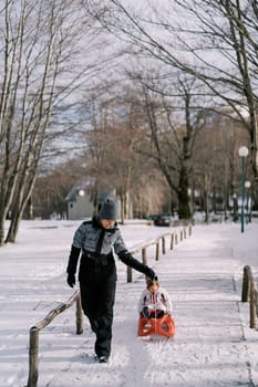 Mom carries a small child on a sled along a snowy path in the park. High quality photo