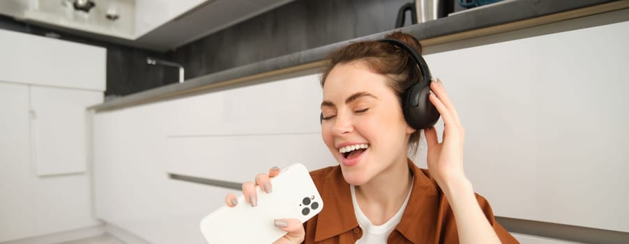 Happy woman listens to music, sings karaoke on smartphone app, sits on floor at home.