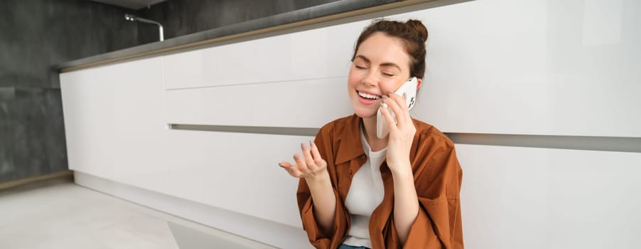 Carefree girl laughs, talks on mobile phone, calling friend and having funny conversation, sitting casually on kitchen floor. Communication and people concept