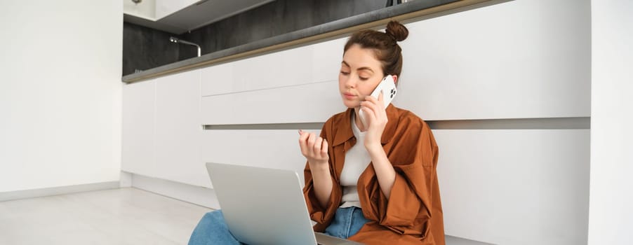 Woman looking bored and unamused while sitting on floor and listening to conversation, has laptop on laps, waiting on line, making phone call.