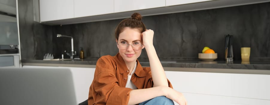 Portrait of young woman, business owner working from home, freelancer using laptop, sitting in kitchen, wearing glasses.