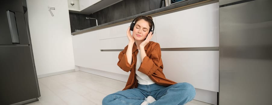 Portrait of young beautiful woman in wireless headphones, sitting on kitchen floor, vibing with favourite music in earphones.