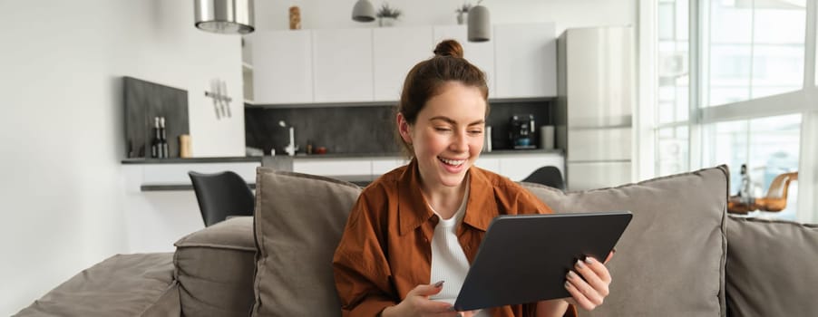 Portrait of young brunette woman reading on digital tablet, watching tv series on her application, sitting on couch in living room.