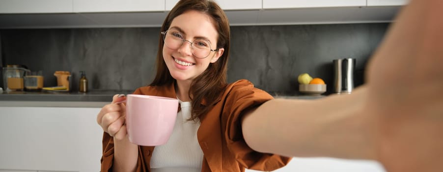 Portrait of beautiful young woman in glasses, taking selfie in kitchen with cup of morning coffee, making photos for her daily routine lifestyle blog.