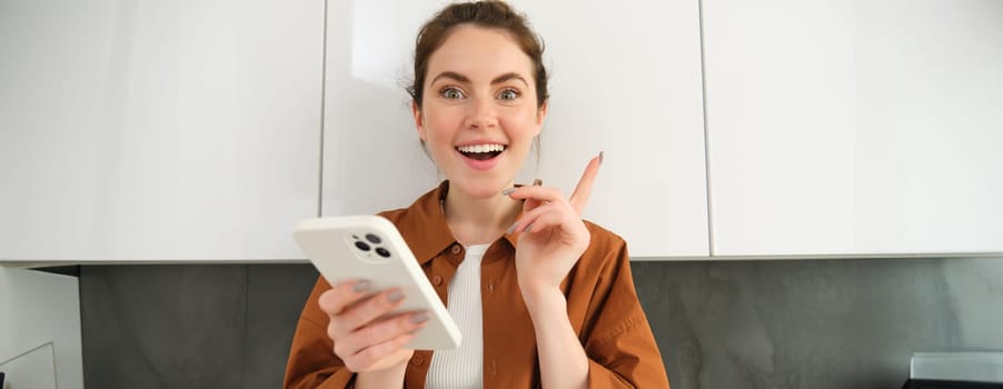Portrait of excited young woman has an idea, holding smartphone and pointing finger up, standing in the kitchen at home.