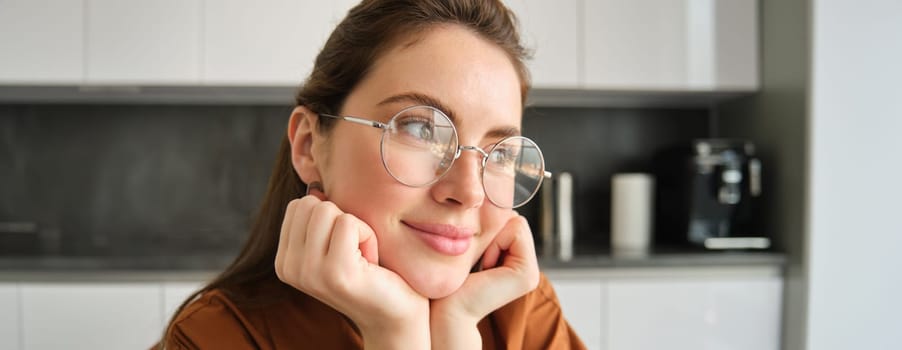 Portrait of lovely, happy young woman, wearing glasses, sitting in kitchen with dreamy, thoughtful face expression, spending time at home.