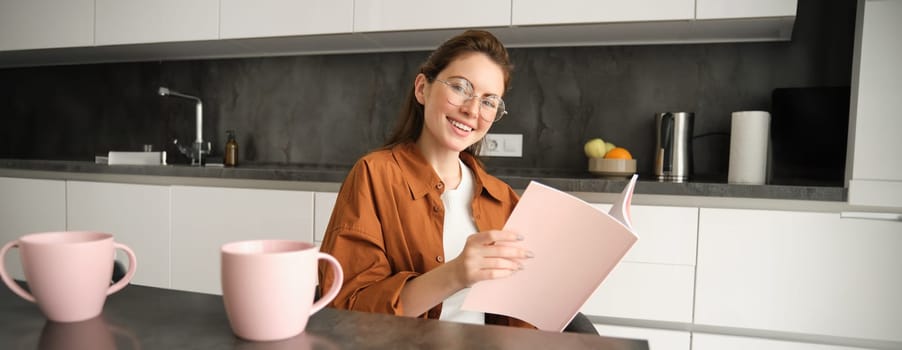 Portrait of young woman working from home, self-employed entrepreneur reading her documents, sitting at home in kitchen, wearing glasses.