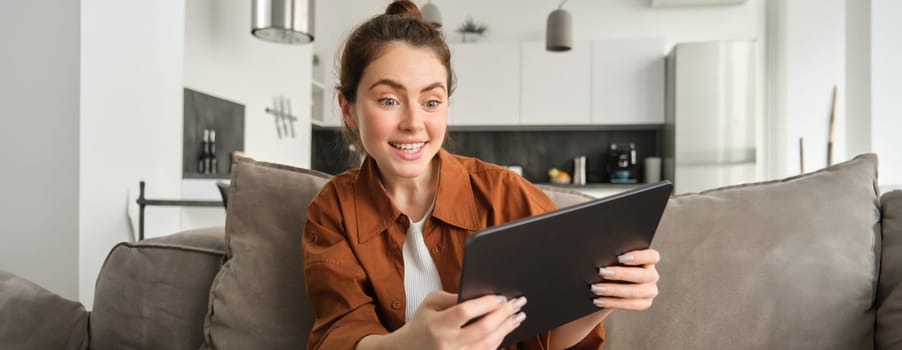 Portrait of excited, smiling young woman playing games on her digital tablet, tilting gadget and looking amused at screen, sitting on couch at home.