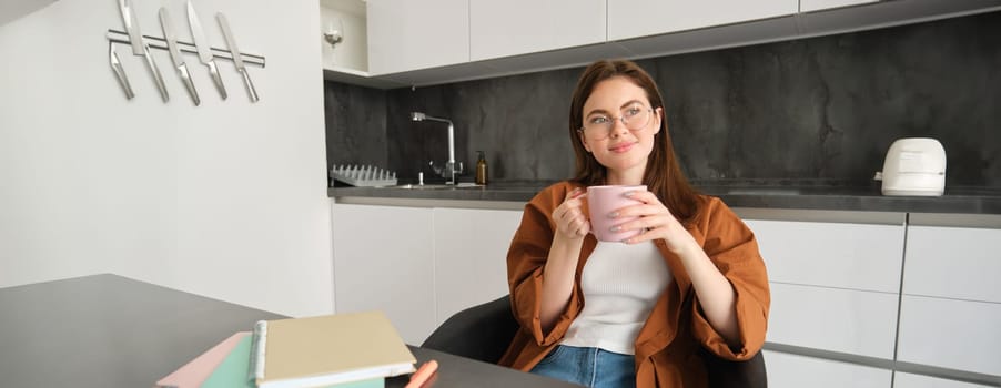 Portrait of smiling young woman in glasses, drinking coffee and looking outside window, sitting in kitchen with homework. Self-employed girl working from home, taking break, enjoying hot tea.
