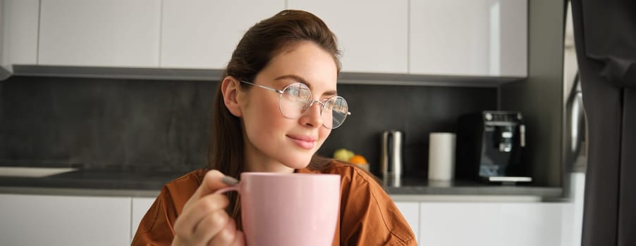 Portrait of beautiful brunette woman, sitting in kitchen, taking a break for cup of coffee. Girl drinks tea at home.