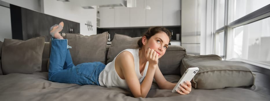 Online shopping concept. Young woman resting on couch, lying on sofa with smartphone, looking up and thinking.