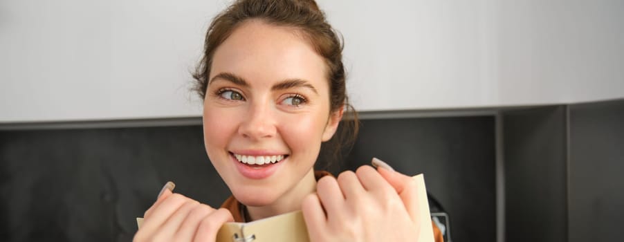 Portrait of young brunette woman in the kitchen, holding notebook, reading notes on planner, laughing and smiling.