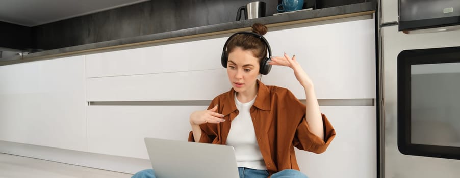 Carefree young woman sits on kitchen floor with laptop, freelancing, digital nomad working from home on computer, listening to music.