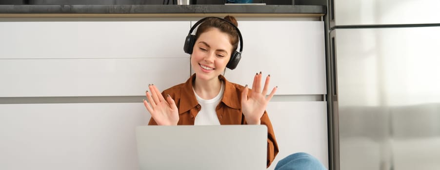 Joyful young woman in headphones, waves hands at laptop, joins online chat, connects to remote course or video call, sits on floor at home in the kitchen.