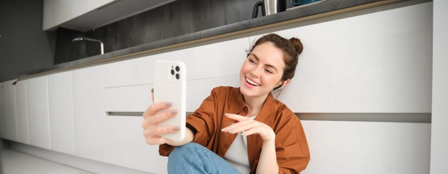 Portrait of beautiful smiling woman, talking to friend on smartphone video chat, connects to online conversation on mobile app, sitting on kitchen floor.