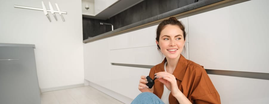 Image of modern brunette girl, sits on kitchen floor, listens to music in wireless headphones, holding earphones and case in hands, looking aside.