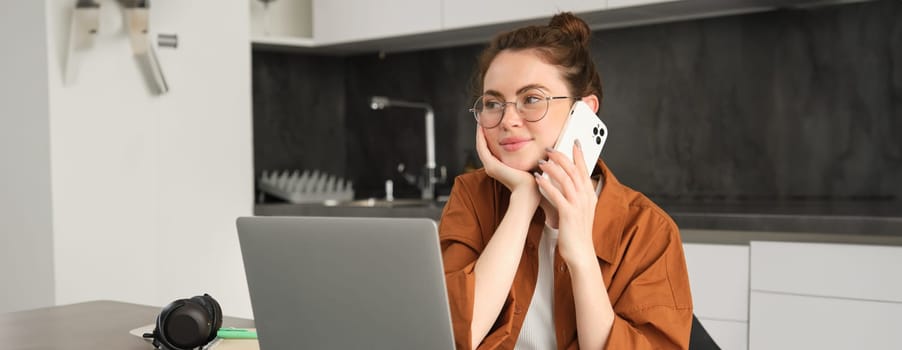 Portrait of young self-employed woman, entrepreneur working from home, freelancer calling client. Girl making an order, talking to someone on phone, sitting in kitchen with laptop.