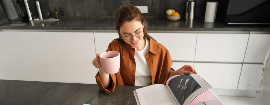 Portrait of beautiful young woman in kitchen, sitting with a book, flipping pages, reading and drinking coffee.