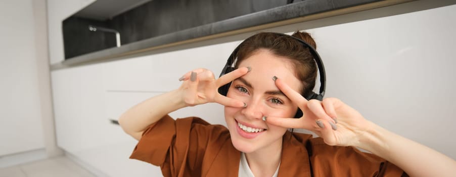 Close up of carefree young modern woman, relaxes at home, listens music in wireless headphones, shows v-sign and smiles at camera.