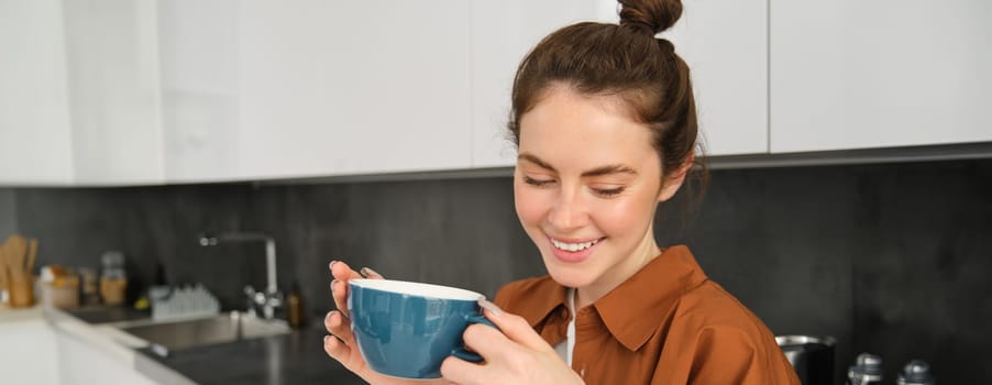 Portrait of beautiful young woman, enjoying delicious aroma of freshly made coffee, holding mug, standing near machine in the kitchen, drinking from mug.