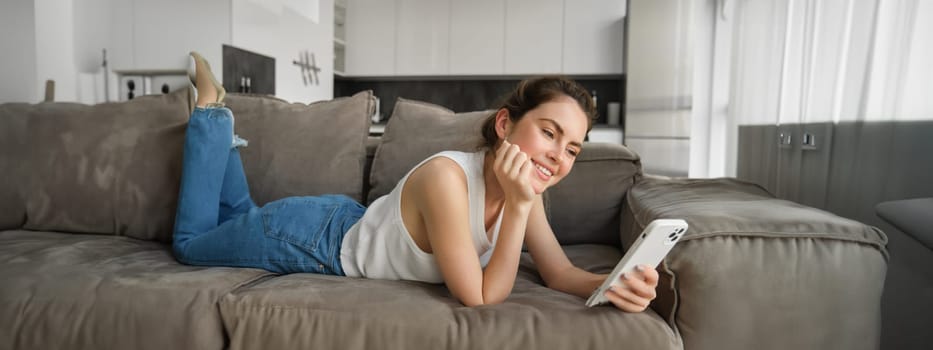 Portrait of cute young woman with smartphone, lying on couch and resting at home, scrolling social media feed, online shopping, reading something on mobile phone.