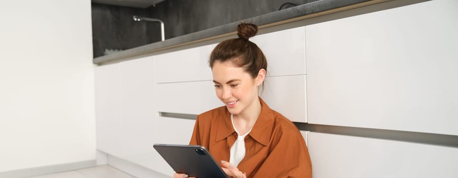 Portrait of young woman working or studying from home, watching her online course on digital tablet, sitting on kitchen floor, smiling and looking happy. People and lifestyle concept