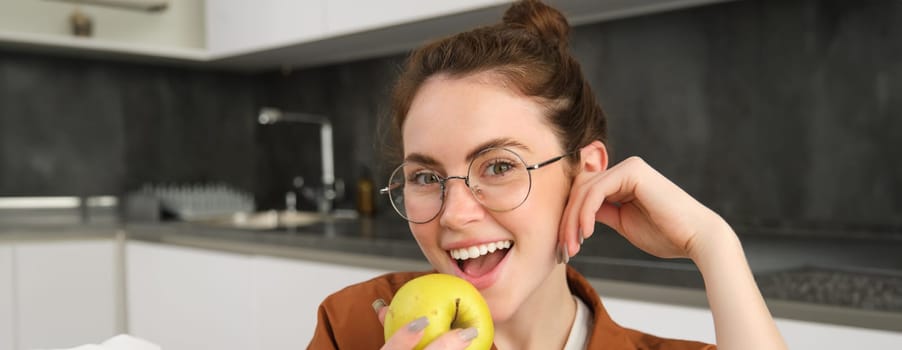 Close up portrait of cute young woman in glasses, eating apple, laughing and smiling, sitting in the kitchen.