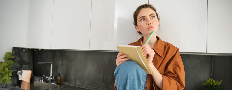 Image of young creative woman, artist drawing sketches in her notebook, sitting on kitchen counter at home with thoughtful face.