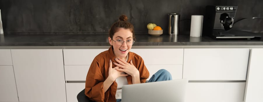Portrait of beautiful woman in glasses, sits at home in kitchen, looks at computer screen with amazed, surprised face, reacts to big great news seeing it on laptop.