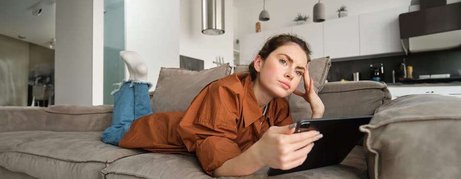 Portrait of woman lying on couch at home, holding digital tablet and looking thoughtful, frowning while thinking and looking aside, making decision, purchase online on gadget.